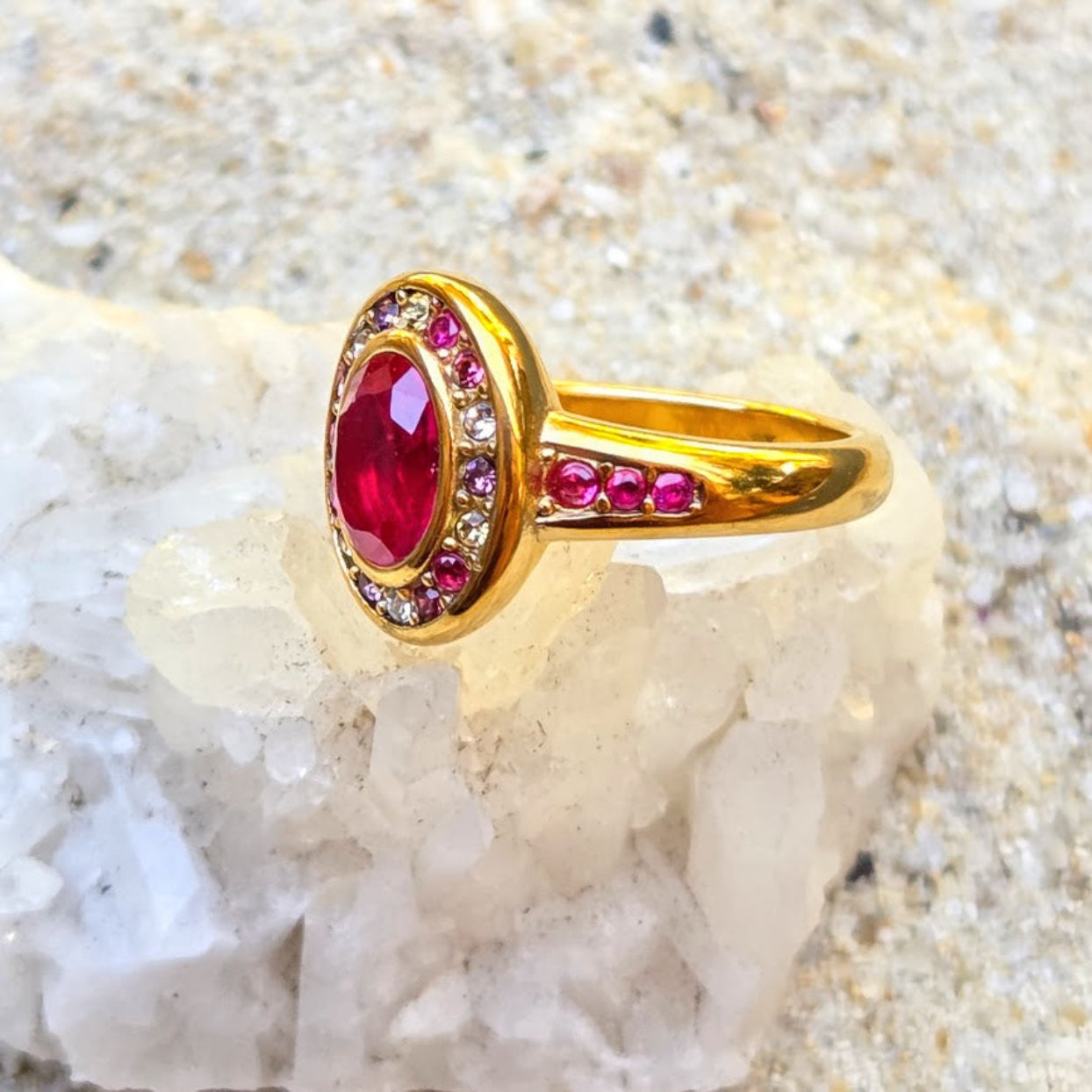 A gold ring with a large oval red gemstone, surrounded by a halo of small clear stones and accented with pink gemstones on the band, displayed on a white crystal against a sandy background.