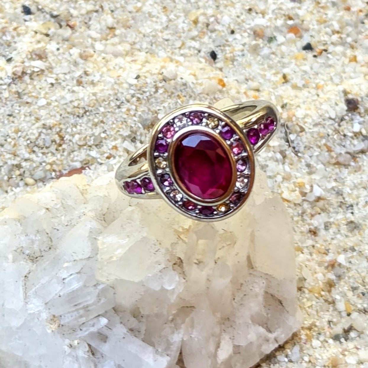 A silver-toned ring with a large oval deep pinkish-red center stone and smaller pink stones around it, displayed on a crystal cluster over sandy ground.