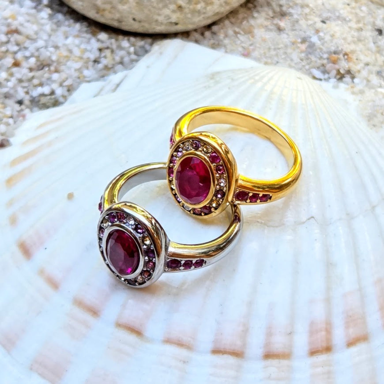 Two gold and silver rings with oval red gemstones and small clear stones, displayed on a white seashell with sand in the background.