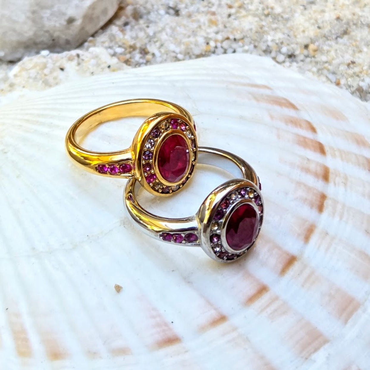 Two gold and silver rings with oval red gemstones and small pink accent stones, displayed on a white seashell with sand in the background.