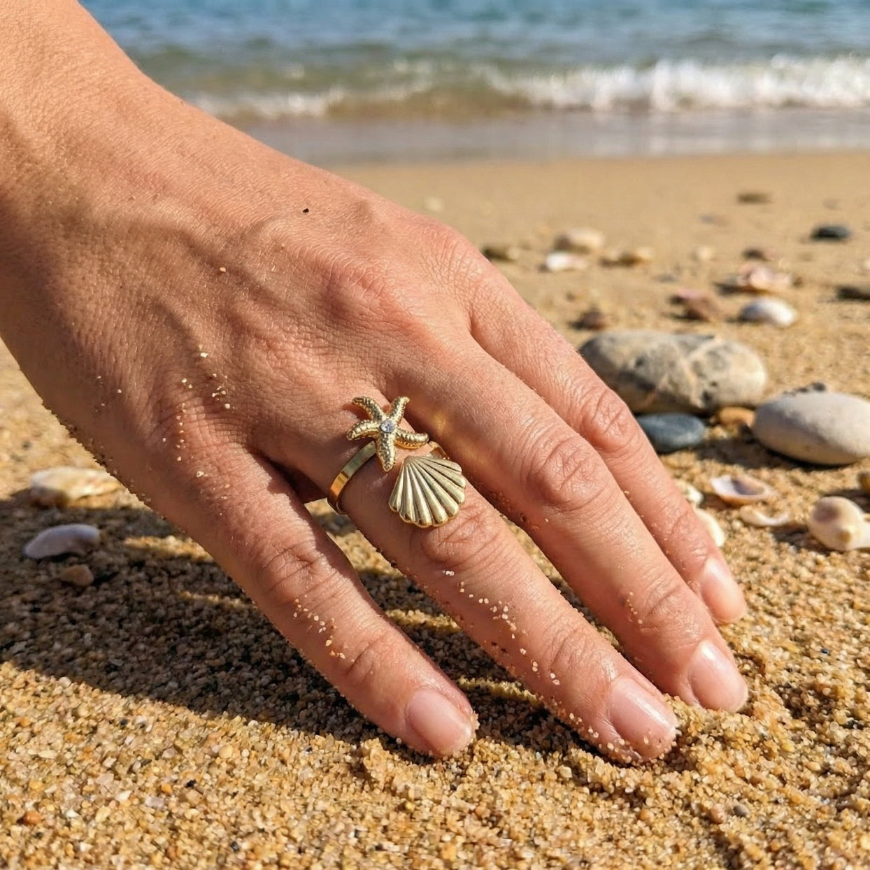 Hand wearing a starfish and shell ring on a sandy beach with ocean waves in the background