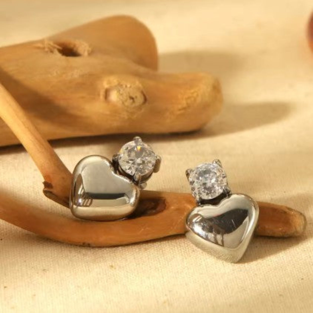 Heart-shaped silver earrings with clear gemstones on a beige background