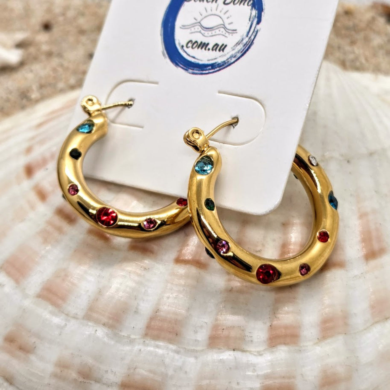 Gold hoop earrings with colorful stones on a shell with a card in the background
