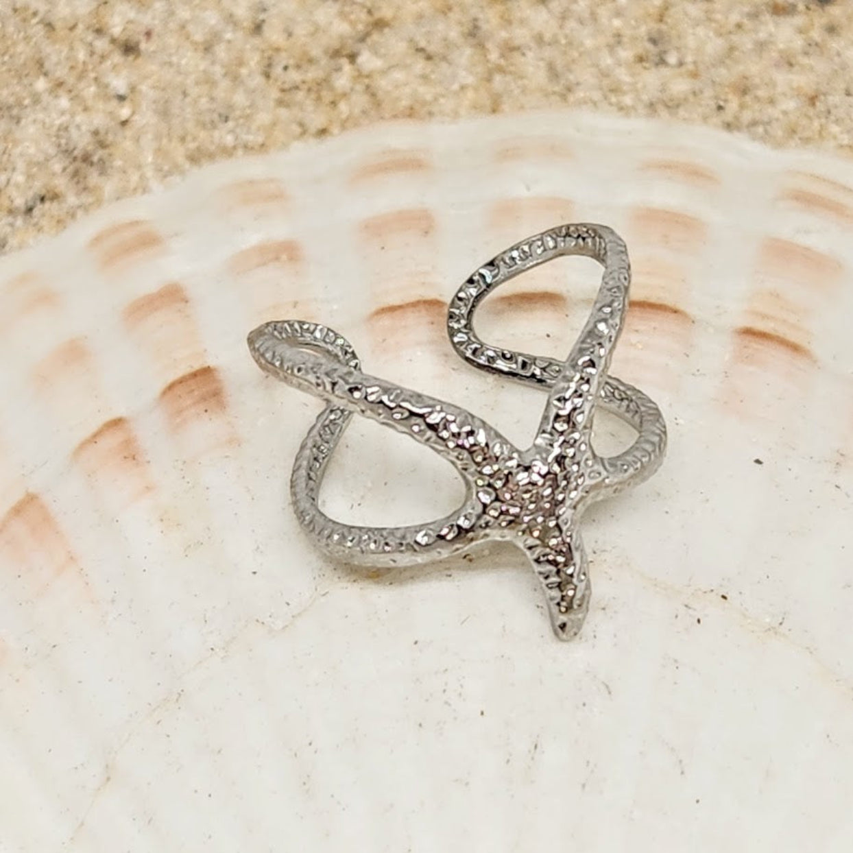Silver starfish-shaped ring on a seashell with sand background