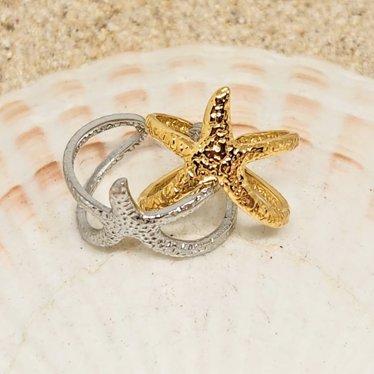 Two starfish-shaped rings, one gold and one silver, on a seashell.