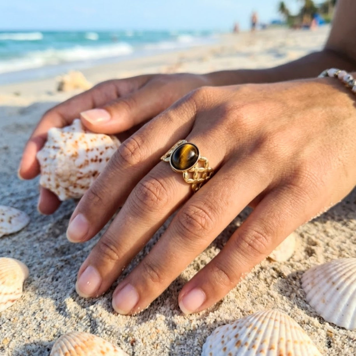 Hand wearing a gold ring with a tiger's eye stone on a sandy beach with shells.