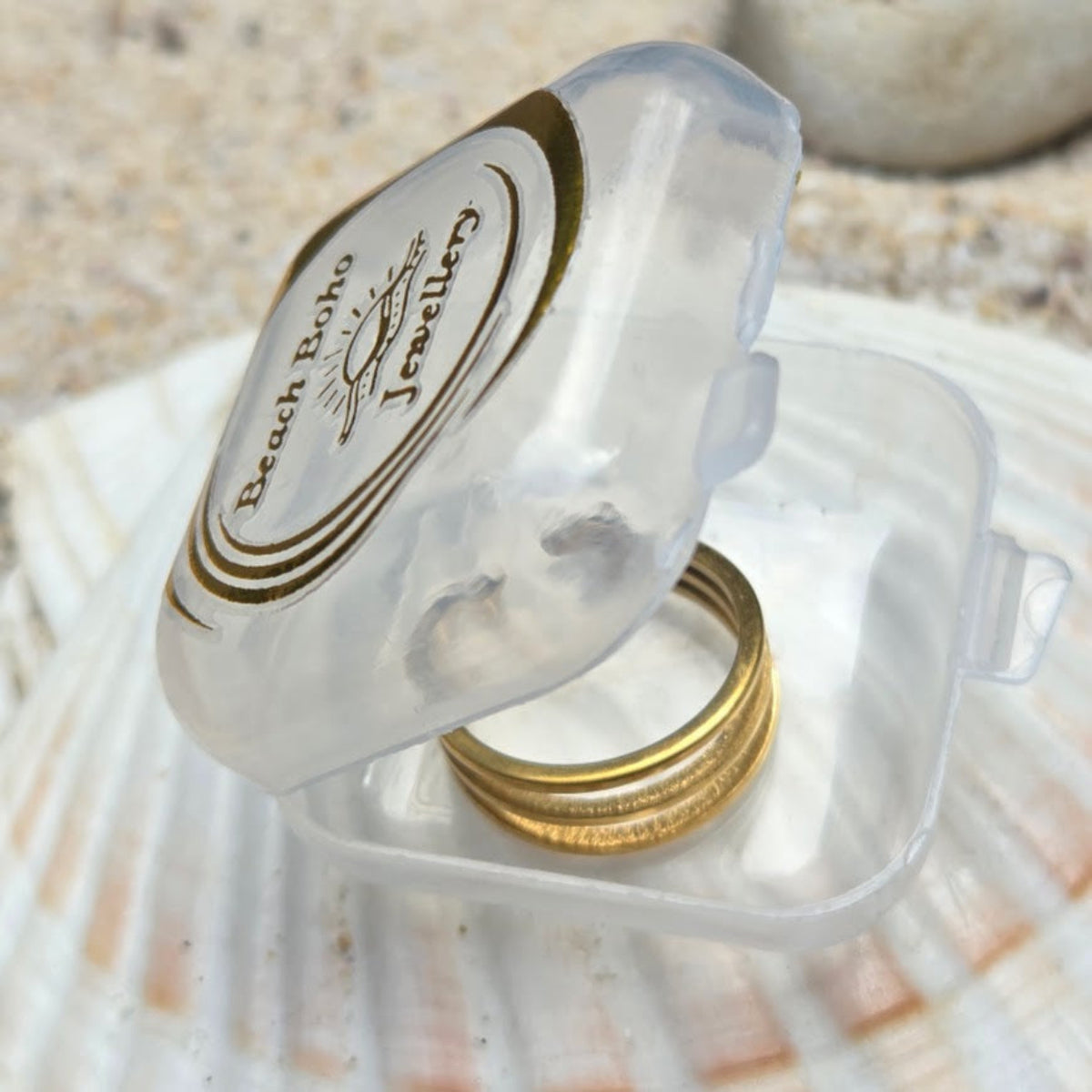 A gold ring displayed inside a clear plastic ring box with a branded lid that reads 'Beach Boats Jewellery', set on a seashell with a sandy background.