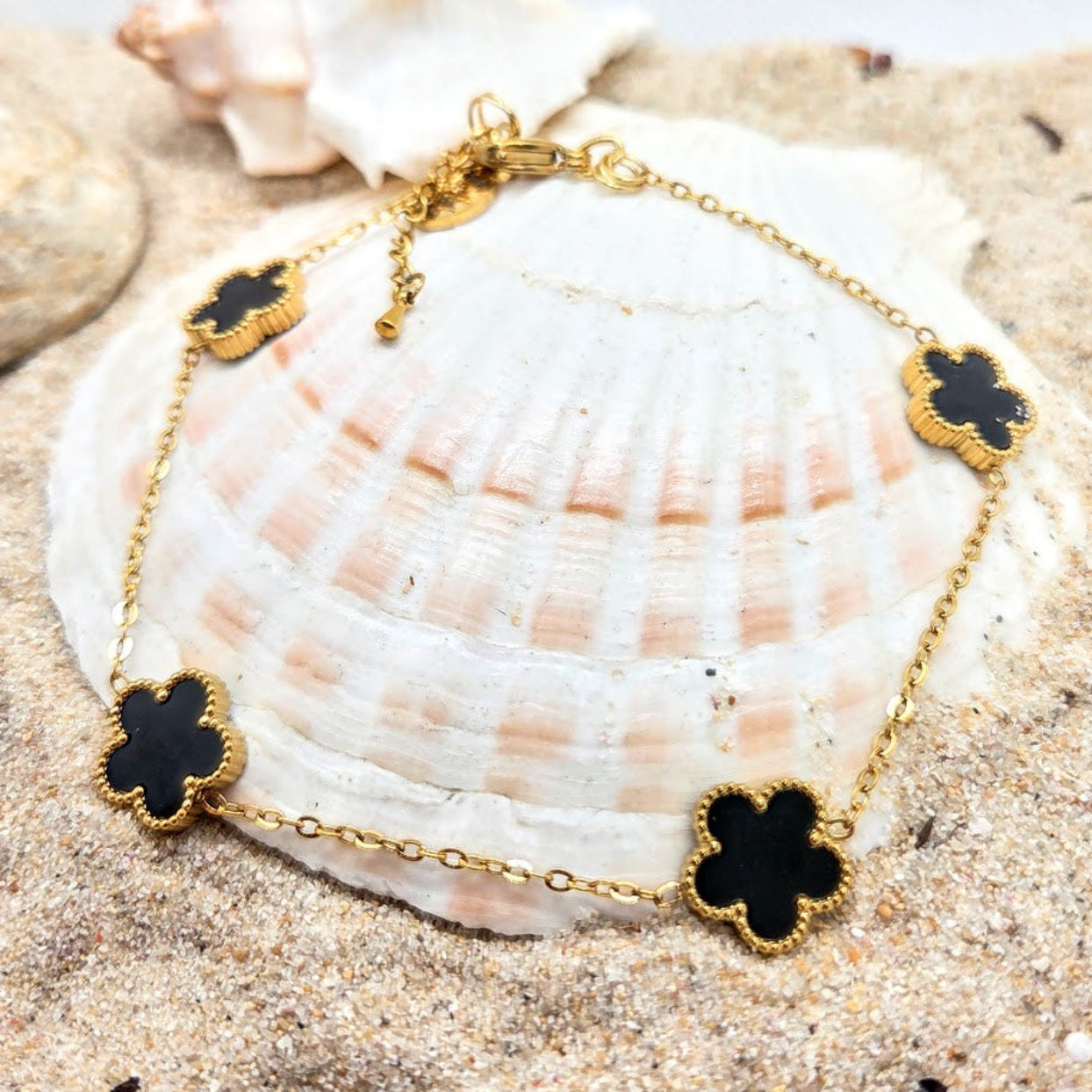 Gold bracelet with black clover-shaped charms on a seashell with sand background